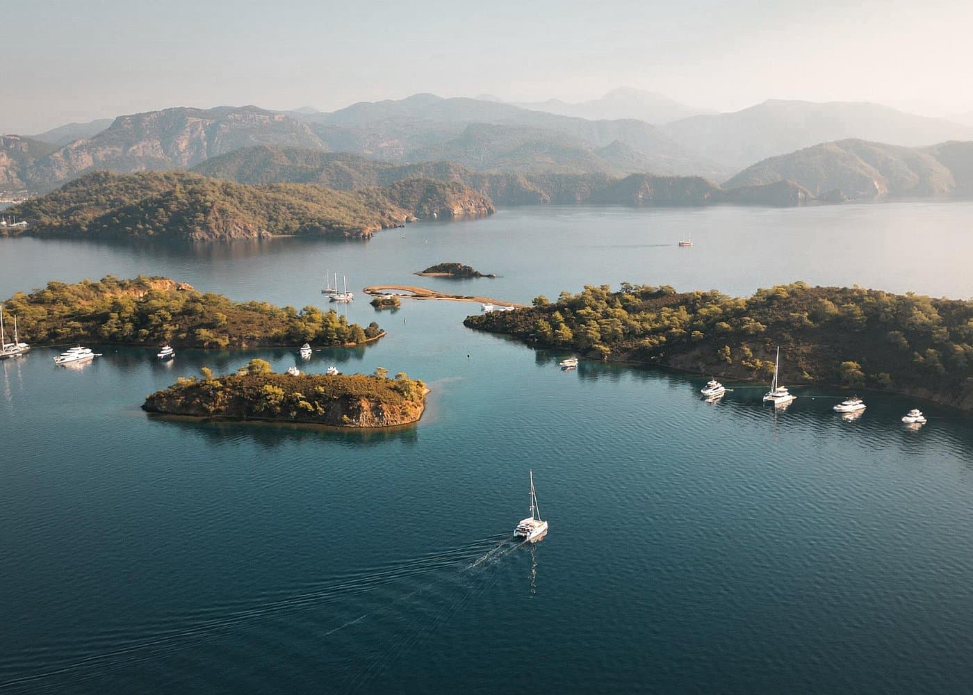 An aerial view of a calm Mediterranean bay with small green islands, moored yachts, and a mountainous coastline