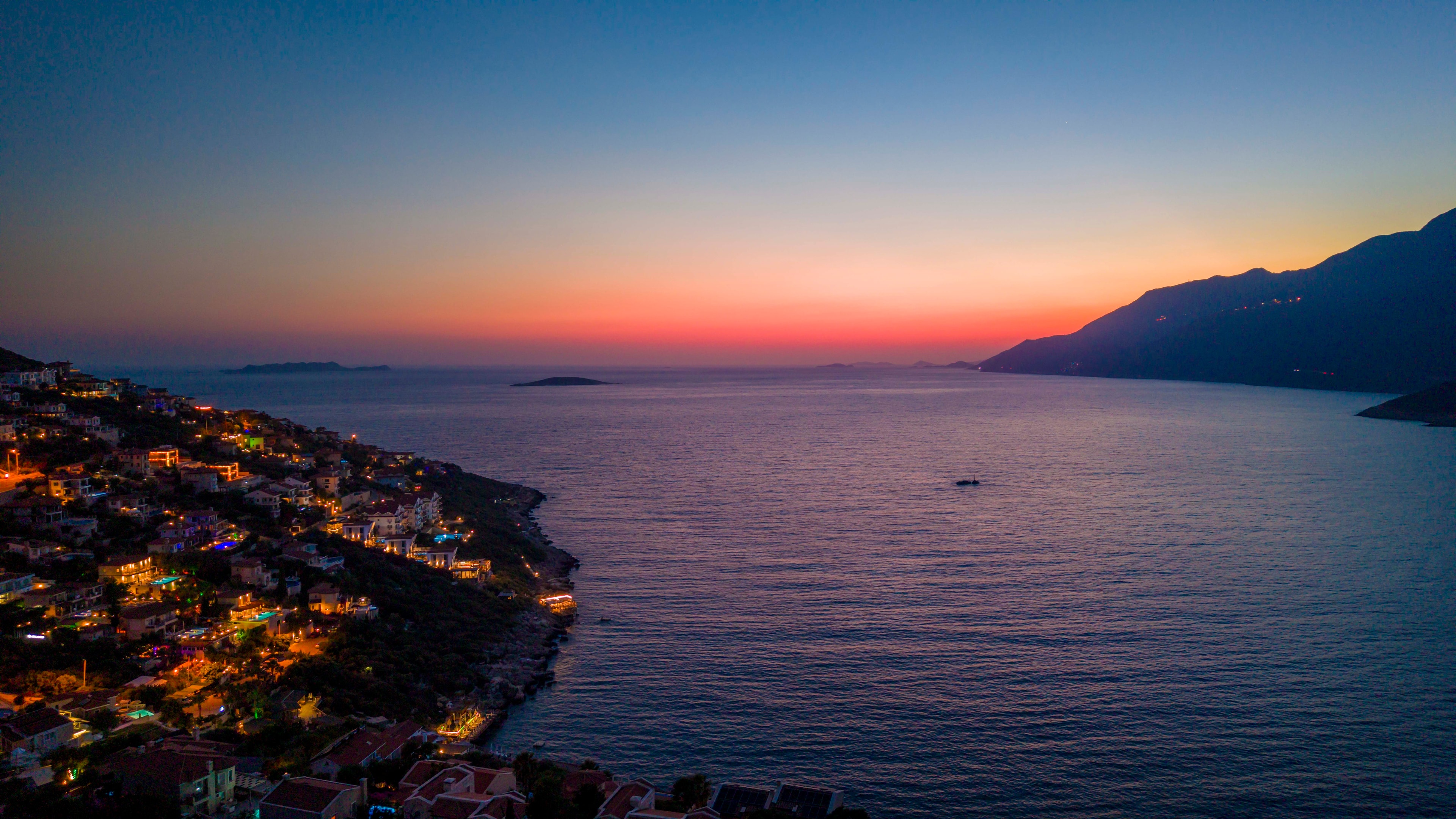 An aerial view of Kalkan's illuminated coastline at dusk, with the open sea and sunset horizon beyond