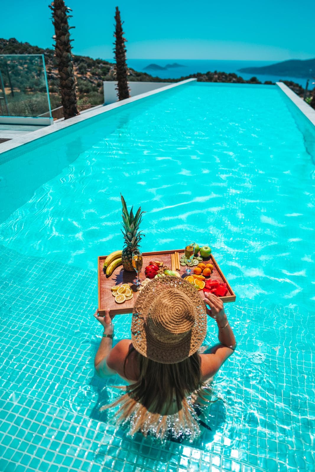 A woman in an infinity pool with a floating breakfast tray, overlooking the Mediterranean coastline